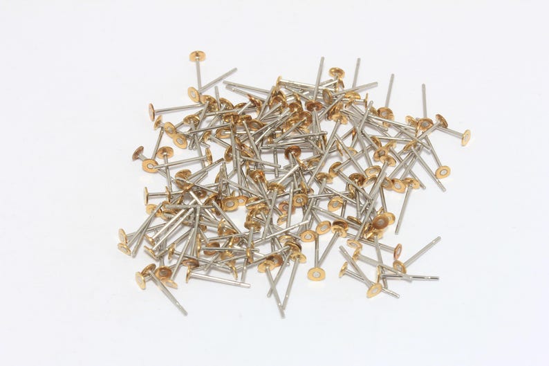 Op de afbeelding: Een stapel kleine, zilverkleurige metalen bevestigingsmiddelen met goudkleurige, platte, cirkelvormige koppen. De bevestigingsmiddelen hebben een lange, dunne, rechte pen. De objecten zijn verspreid op een witte achtergrond.