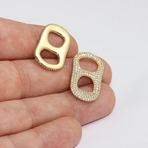 May include: Two gold-colored, rectangular-shaped charms. One charm is solid gold, while the other is encrusted with small, clear crystals. The charms are held in a person's hand against a white background. These charms are likely for jewelry making.