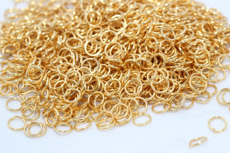 May include: A close-up of a collection of small, gold-coloured jump rings. The rings are circular with a split, enabling them to be opened and closed. They are scattered across a white surface.