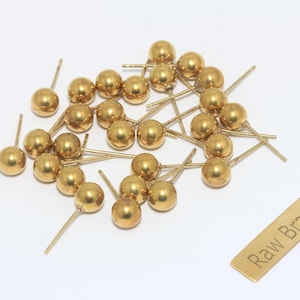 May include: A selection of small, gold-coloured ball stud earrings with thin posts, scattered on a white surface. A small rectangular tag with the text "Raw Brass" is also present. These earrings are ideal for jewelry making.