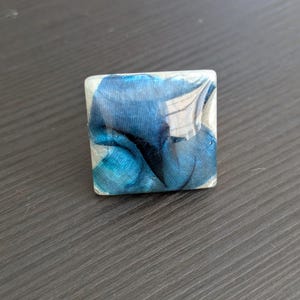 May include: A square-shaped ring with a blue and white abstract design. The ring is made of resin and has a glossy finish.