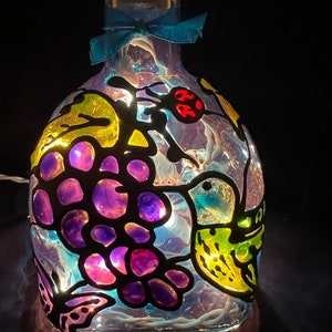 May include: A glass bottle with a blue ribbon around the neck, painted with a hummingbird, grapes, and a ladybug. The bottle is illuminated from within, creating a colorful glow.