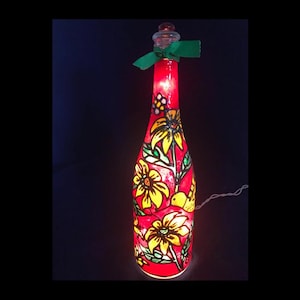 May include: A clear glass bottle illuminated from within by a red light, featuring hand-painted yellow sunflowers with green leaves and stems. A small green ribbon is tied around the neck of the bottle, which is topped with a clear glass stopper.