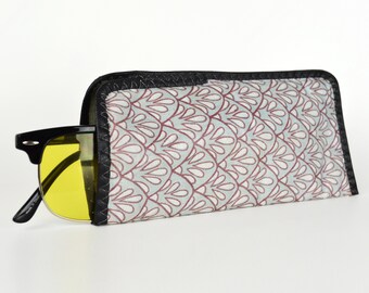 May include: A black and white patterned fabric sunglasses case with a black trim. The case is closed and a pair of yellow-tinted sunglasses are partially sticking out.