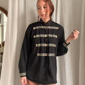 May include: Black long-sleeve shirt with gold horizontal stripes and gold buttons. The shirt has a high collar and gold trim on the cuffs. The shirt is buttoned up the front.