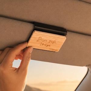 Peut inclure: Un clip rectangulaire en bois clair fixé sur un pare-soleil de voiture. Le bois présente le texte gravé "Drive safe I love you" suivi du nom "James" dans une police cursive. Le clip est fixé sur le pare-soleil en tissu.