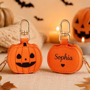 May include: Two orange pumpkin-shaped pouches with gold-tone metal lobster clasps. One pouch features a black jack-o'-lantern face, while the other displays the name Sophia in black script text above a small black heart icon.