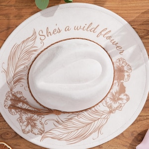 Peut inclure: Un chapeau de cowboy blanc avec un bandeau en cuir marron et un motif d&eacute;coratif de plumes et de fleurs. Le chapeau porte l'inscription "She's a wild flower" sur le bord. Un objet rose en forme de c&oelig;ur avec le texte "She believed she could so she did" est &agrave; c&ocirc;t&eacute; du chapeau.