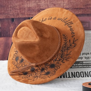 May include: A brown suede cowboy hat with a black floral design and the text "She is a wildflower" around the brim.