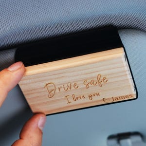 Puede incluir: Un clip de visera de coche de madera con el texto grabado "Drive safe I love you James". El clip es rectangular con un respaldo negro y se sostiene contra una visera de coche.