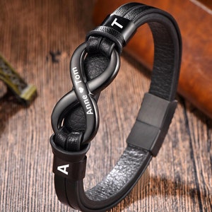 Könnte beinhalten: Schwarzes Lederarmband mit einem Unendlichkeits-Anhänger. Der Anhänger ist mit den Namen "Anne" und "Tom" sowie einem Herzsymbol graviert. Das Armband hat einen schwarzen Metallverschluss und ist an beiden Enden mit den Buchstaben "A" und "T" verziert.
