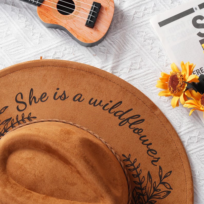 May include: A brown suede hat with a wide brim and a black inscription that reads "She is a wildflower". The hat is decorated with black floral designs.