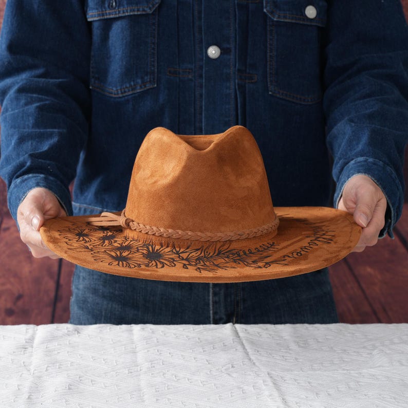 May include: A brown suede cowboy hat with a black sunflower design and a braided band around the crown.