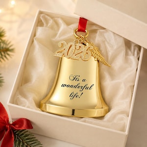 Pode incluir: Um ornamento de sino na cor dourada com o texto gravado "It's a wonderful life!" e um pingente pendurado com o ano "2026" e uma pequena asa de anjo. O sino est&aacute; suspenso por uma fita vermelha e apresentado dentro de uma caixa de presente branca.