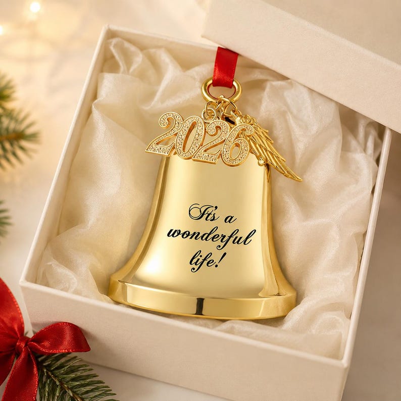 Pode incluir: Um ornamento de sino na cor dourada com o texto gravado "It's a wonderful life!" e um pingente pendurado com o ano "2026" e uma pequena asa de anjo. O sino est&aacute; suspenso por uma fita vermelha e apresentado dentro de uma caixa de presente branca.