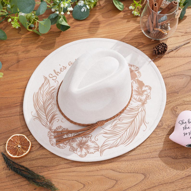 Peut inclure: Un chapeau fedora blanc avec un bandeau en cuir marron et une pampille, pos&eacute; sur une base de chapeau cr&egrave;me. La base pr&eacute;sente un motif floral et de plumes de couleur marron clair, avec le texte "She's a..." imprim&eacute; sur le c&ocirc;t&eacute;.