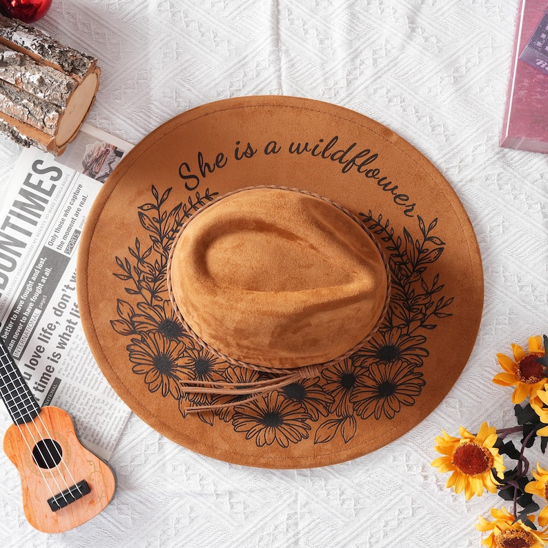 May include: A brown suede hat with a black floral design and the text "She is a wildflower" around the brim.