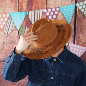 May include: A brown suede cowboy hat with a black floral design and the text "She is a wildflower" written on the brim.