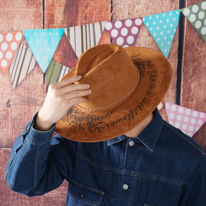May include: A brown suede cowboy hat with a black floral design and the text "She is a wildflower" written on the brim.