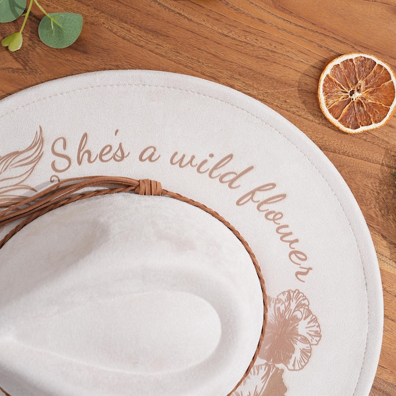 Peut inclure: Chapeau fedora cr&egrave;me avec l'inscription "She's a wild flower" en script marron. Le chapeau est orn&eacute; d'un bandeau en cuir marron et d'un motif d&eacute;coratif de plumes et de fleurs. Une tranche d'orange s&eacute;ch&eacute;e repose sur une surface en bois.