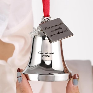 Pode incluir: Um ornamento de sino em metal prateado com uma fita vermelha, um pendente de floco de neve prateado e uma etiqueta quadrada de metal gravada com o texto The sweetest sounds are shared. O sino tem gravado o texto Christmas 2026.