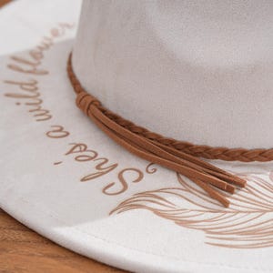 Peut inclure: Chapeau fedora gris clair avec un large bord. Le bord porte l'inscription "She's a wild flower" en marron, ainsi qu'un motif de plume. Un bandeau tress&eacute; marron et des glands ornent le chapeau.