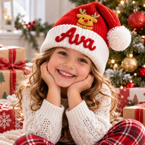 Puede incluir: Un gorro de Navidad de punto rojo y blanco con un pompón blanco en la punta, un borde blanco con el nombre Ava bordado en letra roja y un pequeño reno de peluche sujeto en la parte frontal de la corona roja.