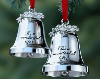 May include: Two polished silver-toned metal bell ornaments hanging from red ribbons. Each bell features a decorative holly leaf topper and the engraved text It's a wonderful life! in a cursive script. The bells have a smooth, reflective surface.