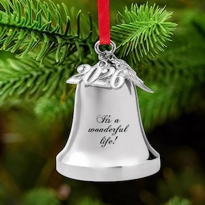 Pode incluir: Um ornamento de sino prateado com o ano 2026 em n&uacute;meros met&aacute;licos em relevo no topo, acompanhado por um pequeno pingente de p&aacute;ssaro. O sino est&aacute; gravado com o texto It's a wonderful life! e est&aacute; suspenso por uma fita vermelha.