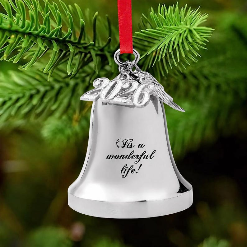 Pode incluir: Um ornamento de sino prateado com o ano 2026 em n&uacute;meros met&aacute;licos em relevo no topo, acompanhado por um pequeno pingente de p&aacute;ssaro. O sino est&aacute; gravado com o texto It's a wonderful life! e est&aacute; suspenso por uma fita vermelha.