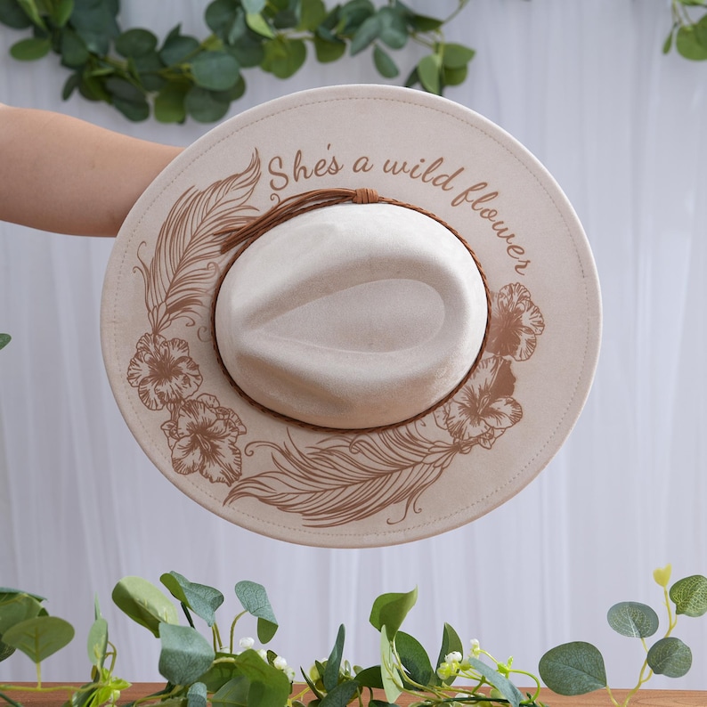 Peut inclure: Chapeau de style fedora cr&egrave;me avec un large bord. Le bord pr&eacute;sente un motif floral et de plumes de couleur marron clair, avec le texte "She's a wild flower" imprim&eacute; sur le bord. Le chapeau a un bandeau marron et une couronne cr&egrave;me.