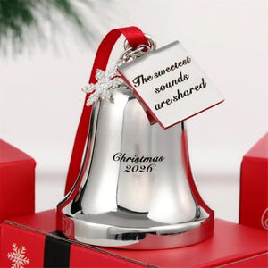 Pode incluir: Um ornamento de sino prateado com uma fita vermelha, um pingente de floco de neve e uma etiqueta quadrada gravada com o texto The sweetest sounds are shared. O sino tem a inscrição Christmas 2026 em uma fonte serifada preta.