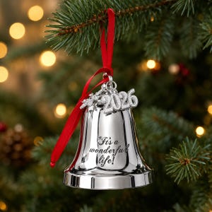Pode incluir: Um ornamento de sino prateado pendurado por uma fita vermelha. O sino apresenta o texto gravado "It's a wonderful life!" e &eacute; adornado com um pingente de floco de neve prateado e um pingente de ano "2026", ambos com acabamento texturizado.