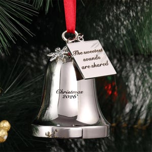 Pode incluir: Um ornamento de sino em metal prateado pendurado por uma fita vermelha. O sino apresenta um pequeno pendente de floco de neve e uma etiqueta quadrada gravada com o texto The sweetest sounds are shared. O sino também tem Christmas 2026.