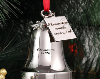 May include: A silver-toned metal bell ornament hanging from a red ribbon. The bell features a small snowflake charm and a square tag engraved with the text The sweetest sounds are shared. The bell is also engraved with the text Christmas 2026.