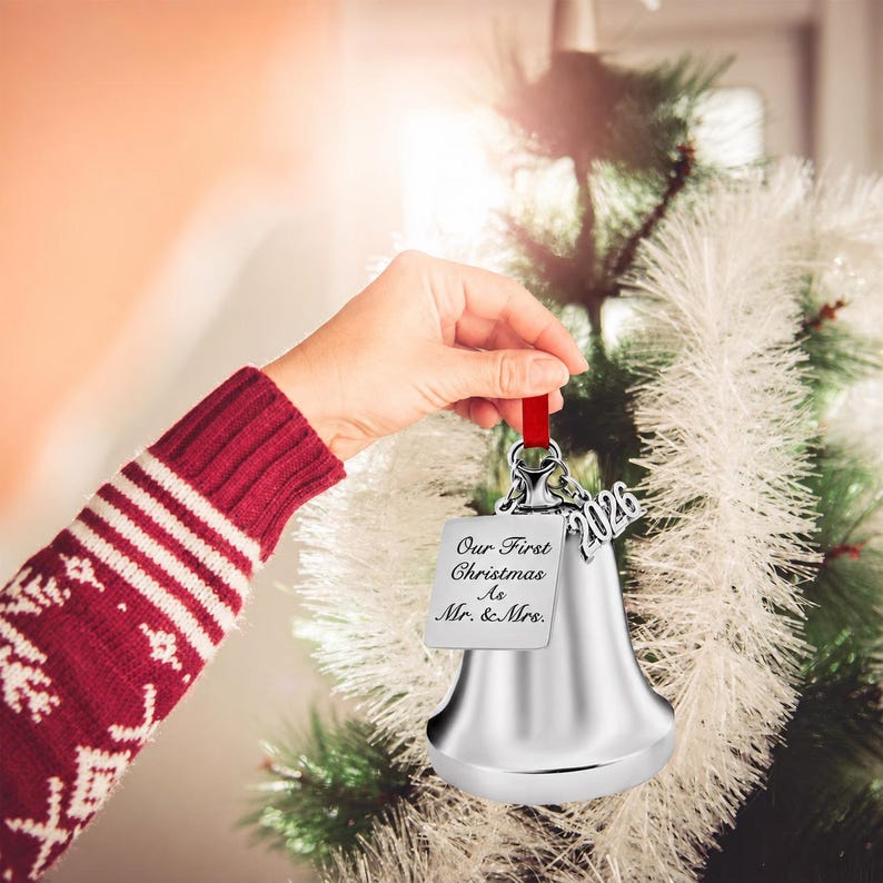 Pode incluir: Um ornamento de sino em tom prateado com uma etiqueta met&aacute;lica quadrada gravada com o texto Our First Christmas As Mr. & Mrs. e um pingente prateado separado exibindo o ano 2026, tudo suspenso por uma fita vermelha para pendurar.
