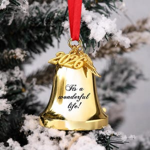Pode incluir: Um ornamento de sino em metal dourado pendurado por uma fita vermelha. O sino apresenta o texto gravado 'It's a wonderful life!' e &eacute; adornado com um pingente dourado que exibe o ano '2026' ao lado de um par de pequenas asas de anjo.