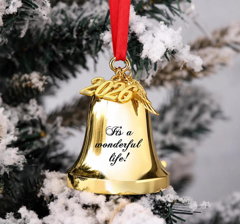 Pode incluir: Um ornamento de sino em metal dourado pendurado por uma fita vermelha. O sino apresenta o texto gravado 'It's a wonderful life!' e &eacute; adornado com um pingente dourado que exibe o ano '2026' ao lado de um par de pequenas asas de anjo.