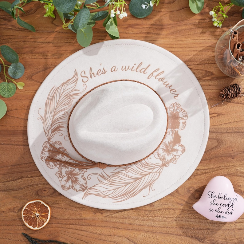 Peut inclure: Un chapeau de cowboy blanc avec un bandeau en cuir marron et un motif d&eacute;coratif de plumes et de fleurs. Le chapeau porte l'inscription "She's a wild flower" sur le bord. Un objet rose en forme de c&oelig;ur avec le texte "She believed she could so she did" est &agrave; c&ocirc;t&eacute; du chapeau.