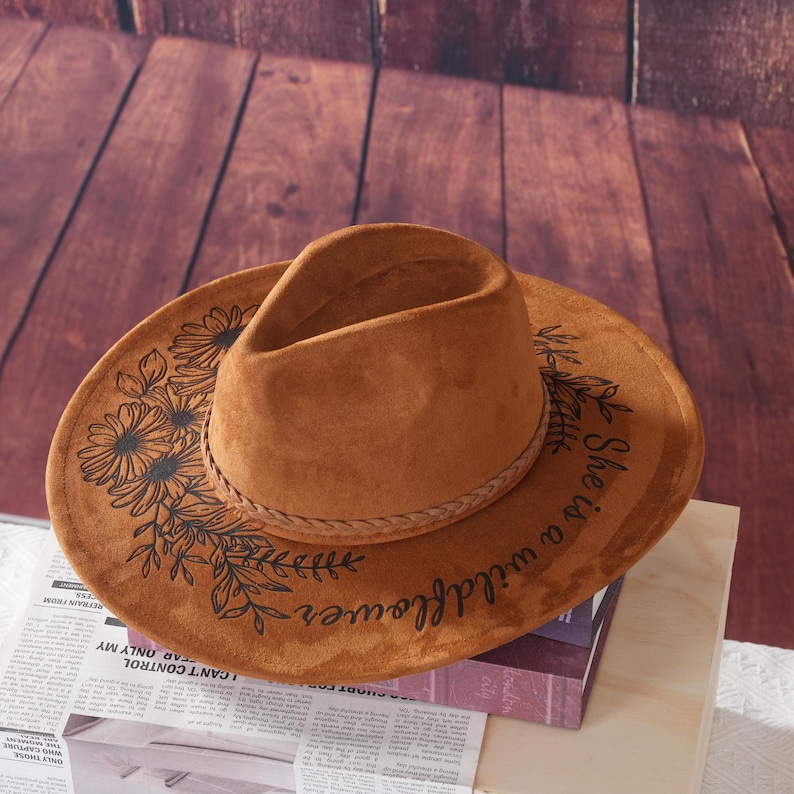 May include: A brown suede cowboy hat with a black braided band. The hat has a floral design and the text "She is a wildflower." printed on the brim.