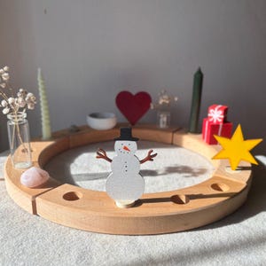 Könnte beinhalten: Ein kreisförmiger Kerzenhalterring aus Holz mit einer weißen Schneemannfigur, die einen schwarzen Hut und eine orangefarbene Nase trägt. Der Ring enthält verschiedene Dekorationselemente wie einen gelben Stern, rote Geschenke, zwei Kerzen und eine Glasvase.