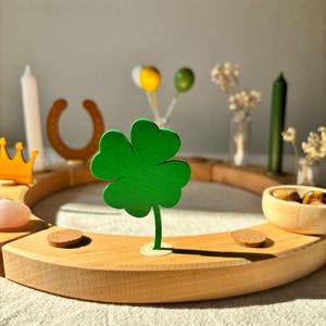 Könnte beinhalten: Ein kreisförmiger Holzring mit einem hellgrünen vierblättrigen Kleeblatt als Einsatz. Das Arrangement umfasst eine weiße Kerze, eine gelbe Krone, ein braunes Hufeisen, eine kleine Holzschale mit Steinen und eine dunkelgrüne Kerze auf dem Holzring.