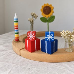 Könnte beinhalten: Ein runder Holzsockel trägt eine gestreifte Kerze, zwei gestapelte rote Geschenkboxen mit weißen Schleifen, zwei gestapelte blaue Geschenkboxen mit weißen Schleifen, eine kleine Glasvase mit Trockenblumen und eine gehäkelte Sonnenblume.