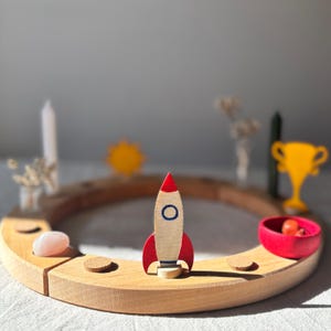 Könnte beinhalten: Ein kreisförmiger Holzring mit einer zentralen Raketenfigur aus Holz, die rote Flügel und ein blaues Bullauge aufweist. Der Ring enthält kleine kreisförmige Vertiefungen, von denen eine einen glatten rosa Stein und eine andere eine rote Schale enthält.