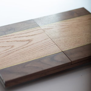 Könnte beinhalten: Ein Schneidebrett aus Holz mit einem Nussbaum- und Eichenholzdesign. Das Brett ist in zwei Abschnitte unterteilt, die jeweils mit einer dünnen Messing-Einlage versehen sind.