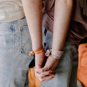 Pode incluir: Duas pessoas usando jeans e camisetas azuis claras, de m&atilde;os dadas. Uma pessoa usa um el&aacute;stico de cabelo laranja, a outra um el&aacute;stico de cabelo rosa. O fundo &eacute; uma superf&iacute;cie laranja.