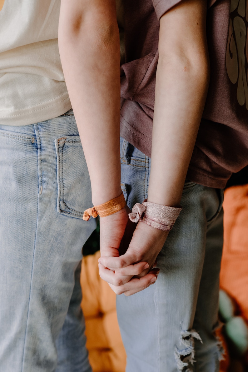 Pode incluir: Duas pessoas usando jeans e camisetas azuis claras, de m&atilde;os dadas. Uma pessoa usa um el&aacute;stico de cabelo laranja, a outra um el&aacute;stico de cabelo rosa. O fundo &eacute; uma superf&iacute;cie laranja.