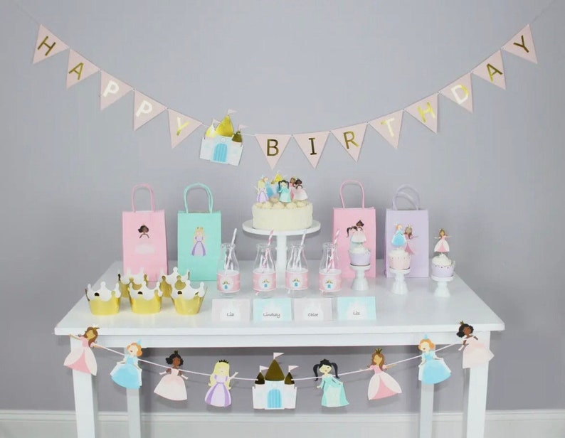 Toddler girls princess party decor in pastel and gold colors.  White table with a cake, gift bags and banner that reads happy birthday with a castle and pastel princess girls.