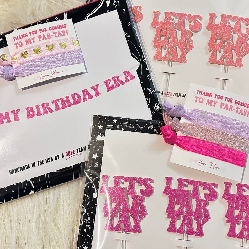 Pode incluir: Uma cole&ccedil;&atilde;o de artigos para festas, incluindo topos de bolo rosa com glitter "Let's Par-tay", el&aacute;sticos de cabelo e uma faixa que diz "My Birthday Era". Os itens s&atilde;o embalados com cart&otilde;es que dizem "Obrigado por vir &agrave; minha festa!" e "Feito &agrave; m&atilde;o nos EUA por uma equipe Dope".
