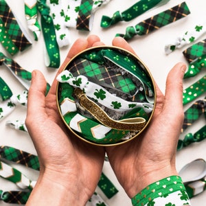 May include: A close-up of a hand holding a container filled with colorful hair ties. The hair ties are made of fabric and have a variety of patterns, including plaid, polka dots, and stripes. The hair ties are arranged in a circular pattern inside the container.